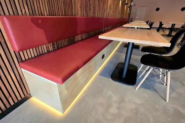 Red leather banquette against the walnut slat wall, lit from below by a warm LED strip, with an oak table and black chairs